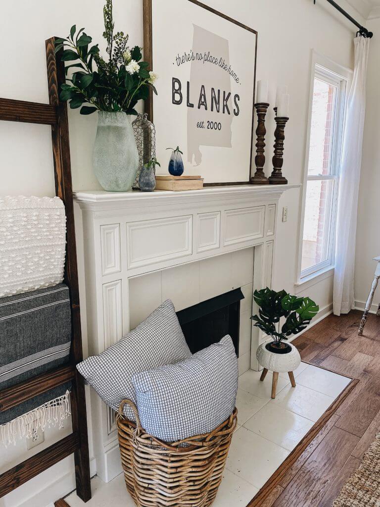 Welcome Home Saturday: Farmhouse fireplace decor items for summer! | Welcome Home Saturday by popular Alabama lifestyle blog, She Gave It A Go: image of a white fireplace decorated with a wooden blanket ladder, woven basket filled with gingham throw pillow, grey plaster vase filled with faux greenery, Alabama state print in a wooden frame, stacked books, and wooden pillar candle stick holders with white pillar candles. 