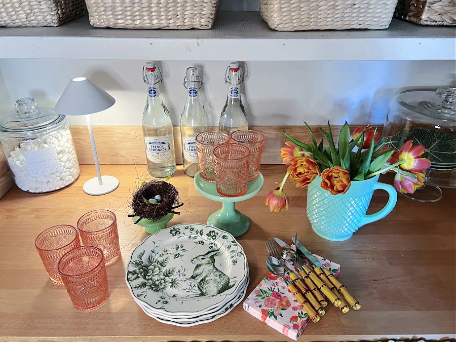 Mini white light, green pitcher with flowers and green and white bunny plates, pink cups and floral napkins on pantry counters.