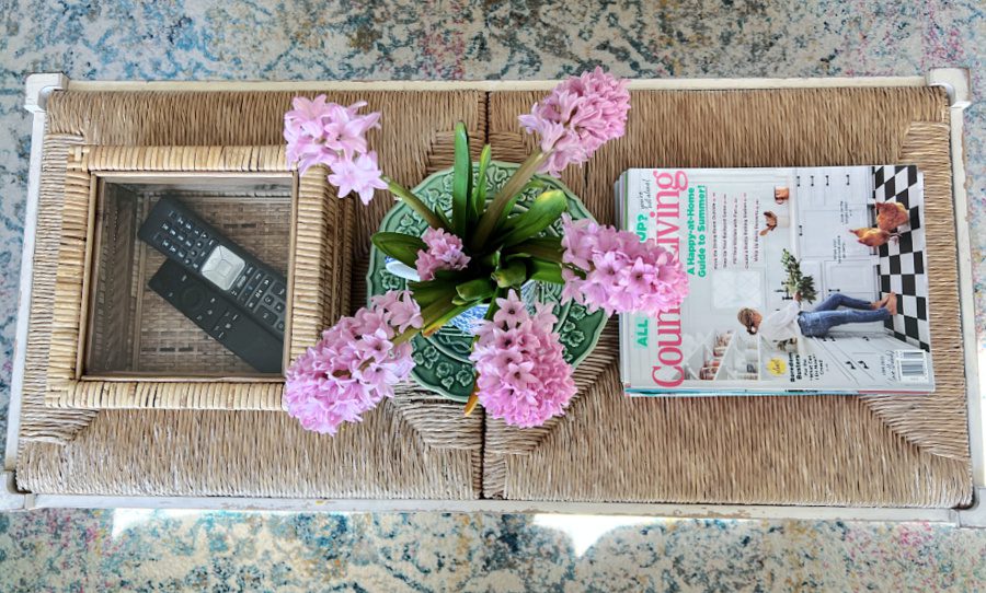 Overhead view of our coffee table with magazines, box holding remotes and a pitcher of pink hyacinths.