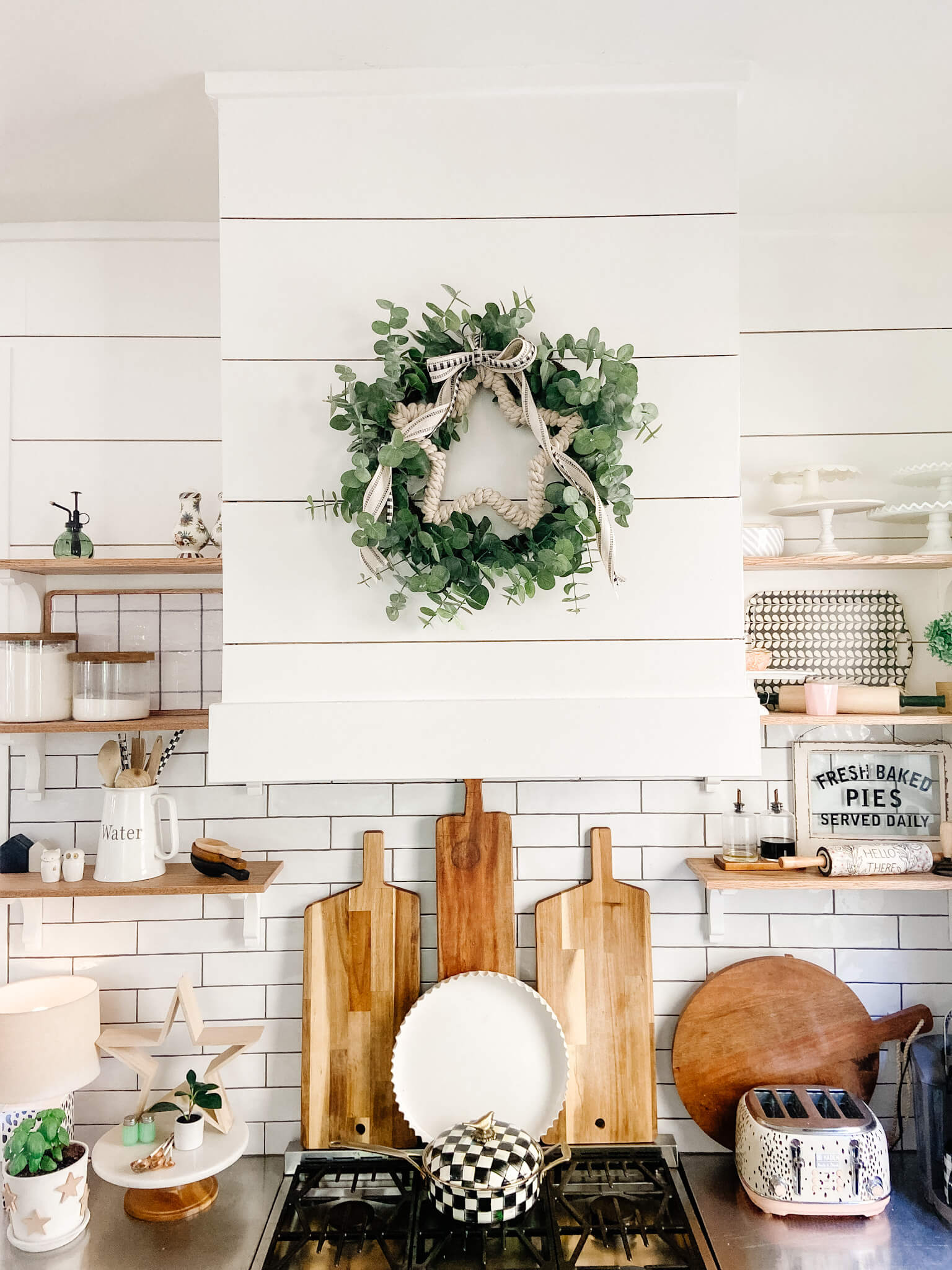 Welcome Home Saturday: Neutral Patriotic Dollar Store Wreath | Welcome Home Saturday by popular Alabama lifestyle blog, She Gave It A Go: image of a farmhouse kitchen with white shiplap walls, white subway tile backdrop, wooden cutting boards, light wood floating shelves decorated with white ceramic dishes, wooden cooking utensils, wire baskets, and filling pins, a gas range stove, and a faux eucalyptus and white pom pom wreath. 