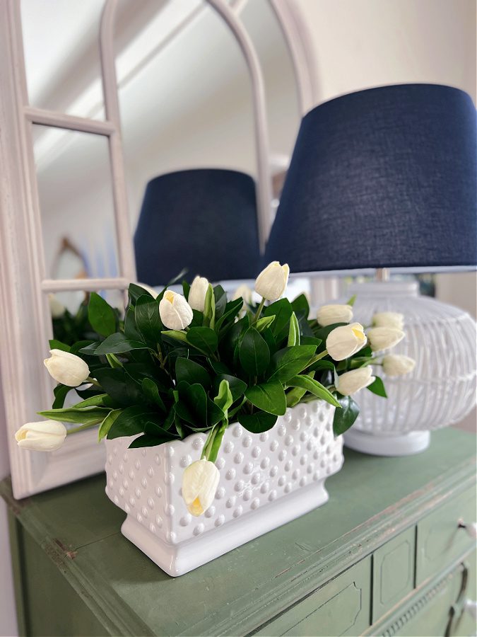 Learn how to make a simple and easy floral arrangement using faux flowers. This tutorial uses tulips, greenery, and a white hobnail planter.