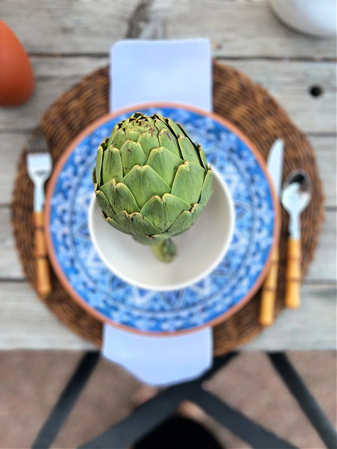 Artichoke in a white bowl on top of melamine dishes and faux bamboo flatware